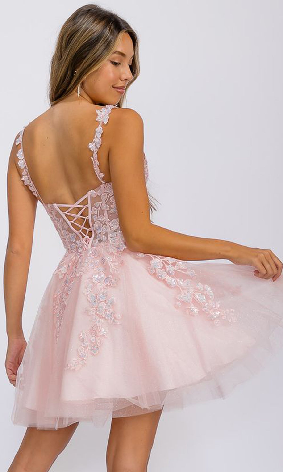 A woman wears the Short Fit-and-Flare Lace Hoco Dress: Eureka EK152, a light pink lace-up party dress with floral lace details and a lace-up back, posing against a plain background.